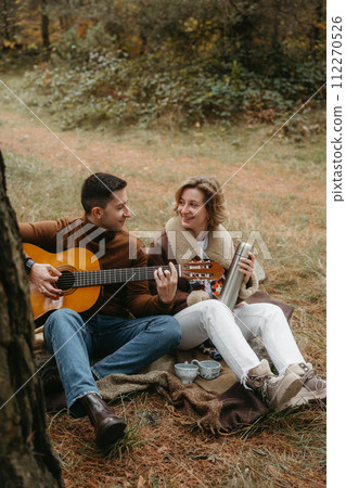 A man is playing guitar for happy woman, adult caucasian couple sitting on blanket in autumn park A man is playing guitar for happy woman, adult caucasian couple sitting on blanket in autumn park 112270526