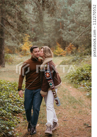 A woman is kissing a man on the cheek while walking through autumn forest, happy couple spending time together 112270552