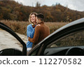 Embracing man and a woman stand next to a car on a dirt road and smiling, couple enjoying roadtrip 112270582