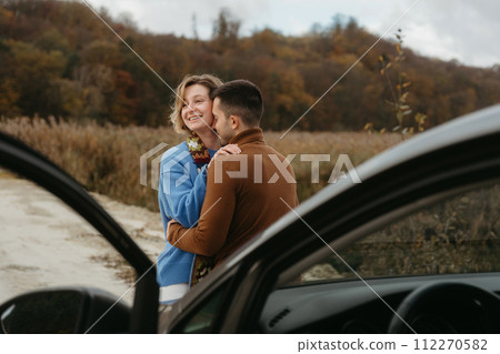 Embracing man and a woman stand next to a car on a dirt road and smiling, couple enjoying roadtrip 112270582