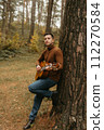 Caucasian adult man plays guitar and leaning on tree in the autumn park 112270584