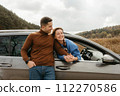 Happy man leaning to a car and holding hands of cheerful woman that sitting inside, adult couple enjoying road trip in autumn 112270586