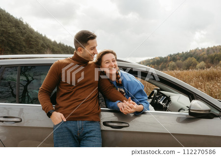 Happy man leaning to a car and holding hands of cheerful woman that sitting inside, adult couple enjoying road trip in autumn Happy man leaning to a car and holding hands of cheerful woman that sitting inside, adult couple enjoying road trip in autumn 112270586
