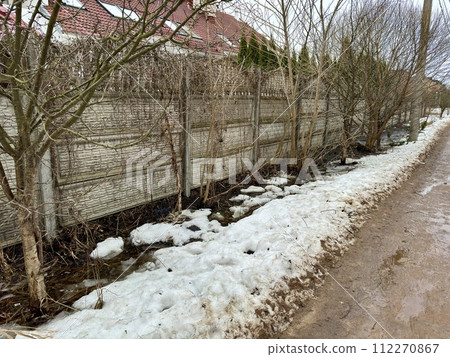Melting dirty snow on the streets of the city during the daytime in winter. The pedestrian road along the fence has snow with sand and mud. Traces of people and pets Melting dirty snow on the streets of the city during the daytime in winter. The pedestrian road along the fence has snow with sand and mud. Traces of people and pets 112270867