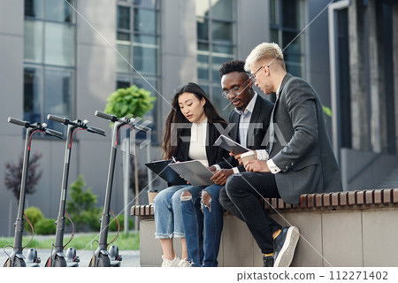 Confident international co workers sitting on wooden bench in front of office and discuss results of joint work. Confident international co workers sitting on wooden bench in front of office and discuss results of joint work. 112271402