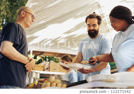 Black woman and caucasian man working together to organize food distribution for hunger relief initiative. Multiracial volunteers serving hot meal from food bank to hungry homeless man. 112271542