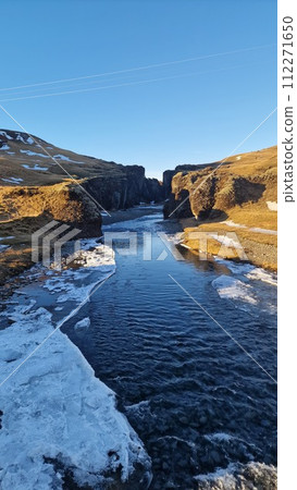 Snowy icelandic canyon with frozen water stream and cliffs covered in frost creating gorgeous landscape view. Magical fjadrargljufur canyon with river flow in iceland between slopes. Snowy icelandic canyon with frozen water stream and cliffs covered in frost creating gorgeous landscape view. Magical fjadrargljufur canyon with river flow in iceland between slopes. 112271650