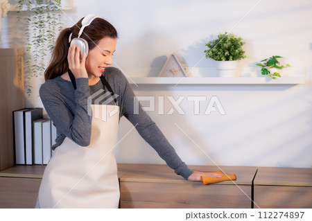 Excited young asian woman with headphones singing and dancing while dusting a wooden cabinet. Excited young asian woman with headphones singing and dancing while dusting a wooden cabinet. 112274897