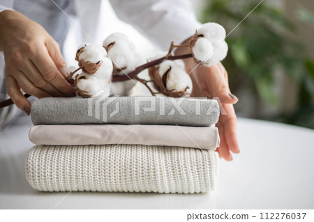 Women's hands hold a stack of neat, clean, cotton clothes in neutral shades. Branch with cotton inflorescences. 112276037