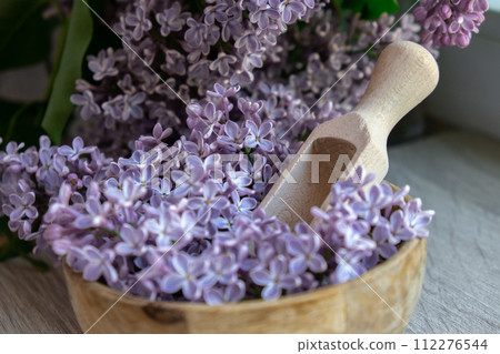 Wooden Bowl with wooden spoon of fresh purple lilac petals with branch of blooming lilac. Lilac flowers fragrance. Concept for spa wellness and aromatherapy. Copy space Still life composition 112276544
