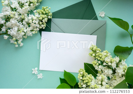 Composition with empty green envelope and beautiful spring lilac flowers on mint background. Mockup card invitation greeting card postcard copy space template. Branches of lilac blooming bouquet.  112276553