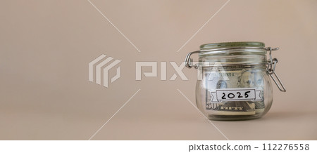 Saving Money In Glass Jar filled with Dollars banknotes. 2025 year transcription in front of jar. Managing personal finances extra income for future insecurity 112276558