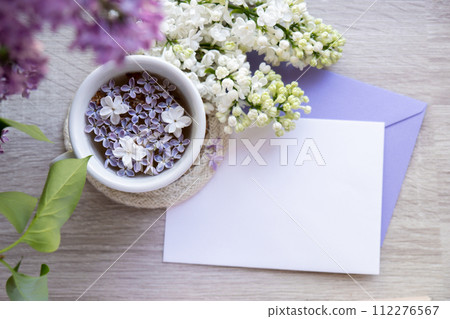 Empty envelope copy space paper note with Tasty black tea in white cup on windowsill with aromatic lilac flowers. Spring composition Cup of lilac tea drinking recipe flowering branches of purple lilac 112276567