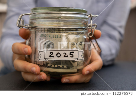 Unrecognizable woman holding Saving Money In Glass Jar filled with Dollars banknotes. 2025 year transcription in front of jar. Managing personal finances extra income for future insecurity 112276573