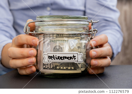 Unrecognizable woman holding Saving Money In Glass Jar filled with Dollars banknotes. RETIREMENT transcription in front of jar. Managing personal finances extra income for future insecurity 112276579