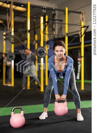 Young dedicated woman doing exercises with kettlebell near other people in crossfit gym Young dedicated woman doing exercises with kettlebell near other people in crossfit gym 112277926