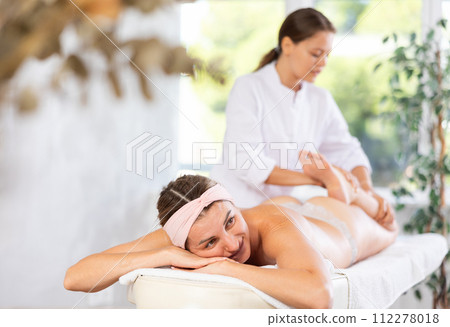 Smiling young woman reclining on massage table while masseuse stretching legs 112278018