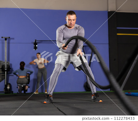 Concentrated man performing battle rope exercise in gym 112278433