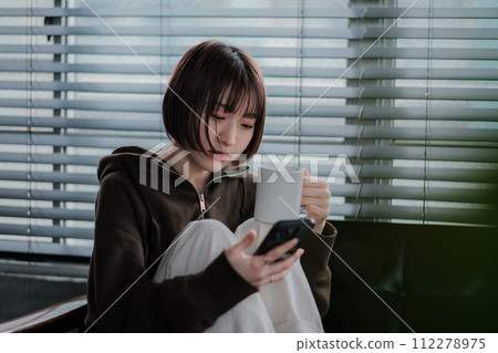 A woman in a loungewear sitting on a sofa and looking at a smartphone A woman in a loungewear sitting on a sofa and looking at a smartphone 112278975