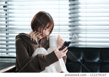 A woman relaxing in her loungewear while looking at her smartphone on the sofa in the morning A woman relaxing in her loungewear while looking at her smartphone on the sofa in the morning 112279107