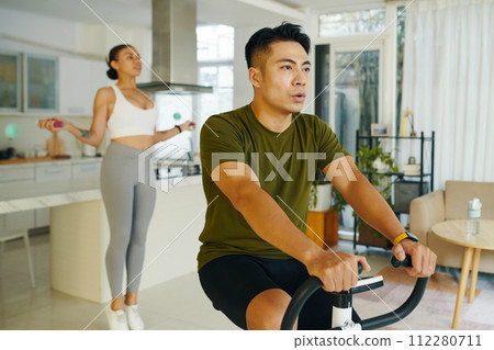 Fit man riding exercise bike when his girlfriend jumping with skipping rope Fit man riding exercise bike when his girlfriend jumping with skipping rope 112280711