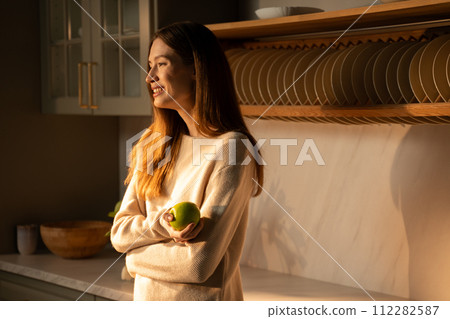 A young woman with an apple in her hands in a sunny modern kitchen at sunrise A young woman with an apple in her hands in a sunny modern kitchen at sunrise 112282587