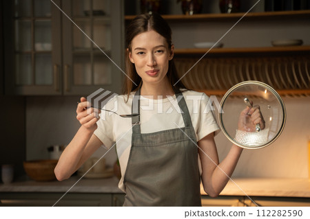 Young Woman in Apron Tasting Soup From a Ladle in a Home Kitchen Setting 112282590