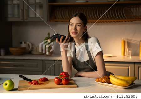 Young Woman Recording a Voice Message in a Modern Kitchen During Meal Preparation 112282605