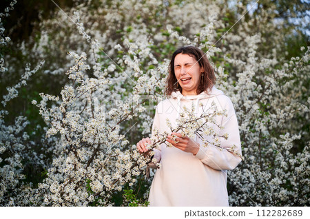 Woman allergic suffering from seasonal allergy at spring, posing in blossoming garden at springtime. Portrait of young woman with eyes closed sneezing among blooming trees. Spring allergy concept Woman allergic suffering from seasonal allergy at spring, posing in blossoming garden at springtime. Portrait of young woman with eyes closed sneezing among blooming trees. Spring allergy concept 112282689