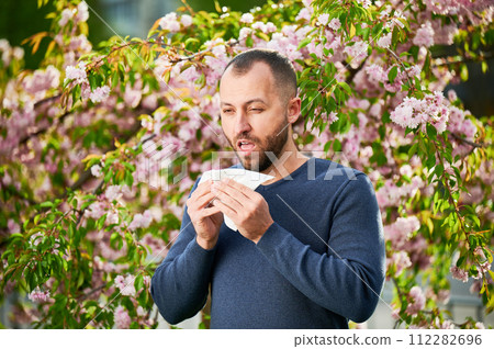 Man allergic suffering from seasonal allergy at spring in blossoming garden at springtime. Man sneezing and blowing nose using nasal handkerchief in front of blooming tree. Spring allergy concept. 112282696