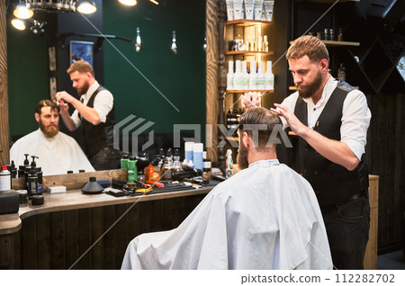 Confident man getting new hairstyle. Experienced barber cutting wet hair. Regular appointment in barbershop. Confident man getting new hairstyle. Experienced barber cutting wet hair. Regular appointment in barbershop. 112282702