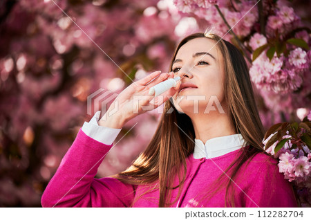 Woman allergic using medical nasal drops, suffering from seasonal allergy at spring in blossoming garden. Close up of woman treating runny nose near blooming tree outdoors. Spring allergy concept. Woman allergic using medical nasal drops, suffering from seasonal allergy at spring in blossoming garden. Close up of woman treating runny nose near blooming tree outdoors. Spring allergy concept. 112282704