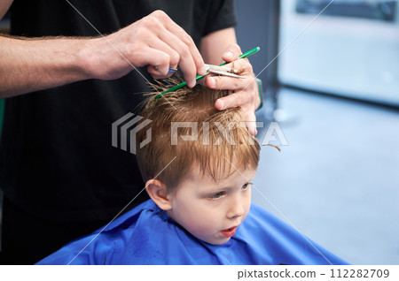 Man barber cutting little boy's hair using comb and scissors. Child getting haircut from adult male, likely barber. Professional hairdresser and cute client at modern barbershop. 112282709