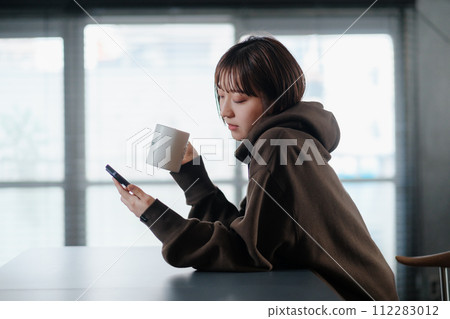 A woman looking at her smartphone while drinking coffee in her room in the morning 112283012