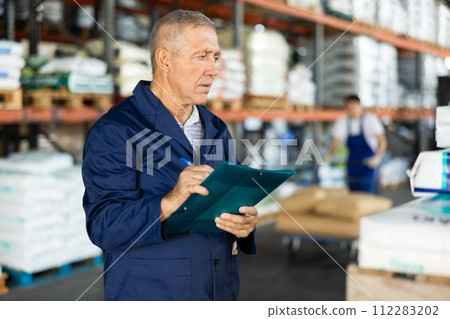 In warehouse of store, mature man checks availability of goods and receipt documents for products 112283202