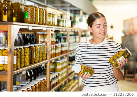 Young female shopper deciding between varieties of pickled olives in store Young female shopper deciding between varieties of pickled olives in store 112283206
