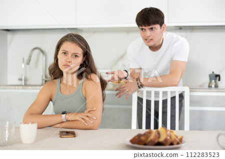 Young girl sitting at the table while her boyfriend is quarreling with her standing behind 112283523