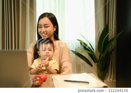 Shot of a young mother having video call with her clients over laptop and carrying her baby on laps 112284238
