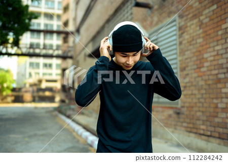Young stylish man listening to music with wireless headphone while walking along a city street 112284242
