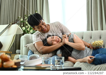 Cheerful Asian couple relaxing on couch and enjoying leisure weekend time at home together Cheerful Asian couple relaxing on couch and enjoying leisure weekend time at home together 112284287