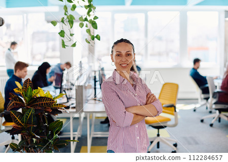 Portrait of young smiling business woman in creative open space coworking startup office. Successful businesswoman standing in office with copyspace. Coworkers working in background 112284657