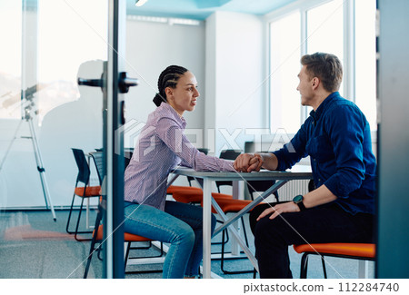 In a modern startup office, a businessman and a businesswoman business colleagues engage in a symbolic arm-wrestling match, reflecting teamwork, competition, and innovation in their dynamic workplace. 112284740