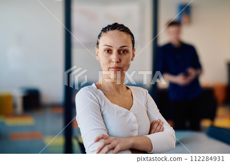 Portrait of a business woman in a creative open space coworking startup office with crossed arms. Successful businesswoman standing in office with copyspace. Associates work in the background 112284931