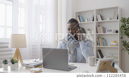 Serious African American male manager is talking on the phone and typing on his laptop, taking an order 112285458