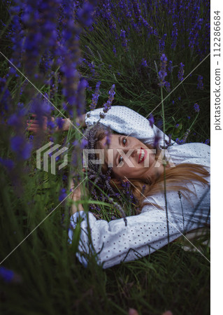 There is a smiling woman in a white dress lying in a lavender field wearing a wreath 112286684