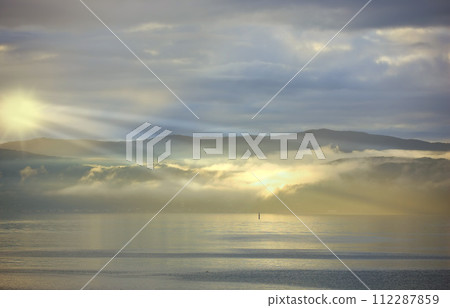 Sea, sunset and cloud on mountains in morning, reflection and island with landscape in nature. Blue sky, clouds or sunlight on water on beach, calm or sunrays on sustainable environment or seascape Sea, sunset and cloud on mountains in morning, reflection and island with landscape in nature. Blue sky, clouds or sunlight on water on beach, calm or sunrays on sustainable environment or seascape 112287859