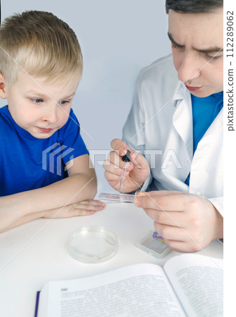 Child chemist. Teacher shows a visual experiment. A science mentor teaches an experimental approach. Microscope, petri dish, pipettes, books. Practical work in chemistry or physics. Laboratory work 112289062
