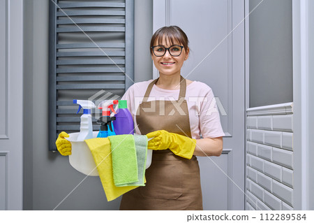 Middle-aged woman in gloves apron with basin of detergents in bathroom Middle-aged woman in gloves apron with basin of detergents in bathroom 112289584