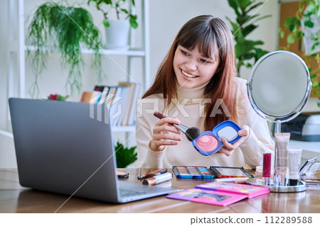 Young female showing cosmetics to laptop webcam, sitting at table at home 112289588