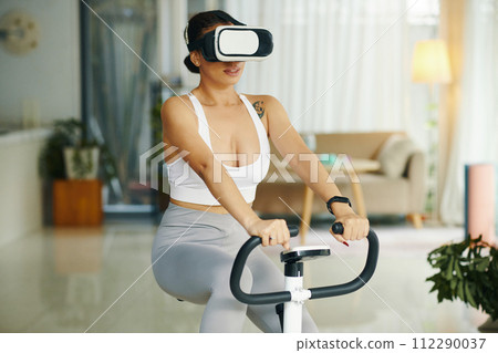 Fit woman wearing virtual reality headset when riding exercise bike Fit woman wearing virtual reality headset when riding exercise bike 112290037